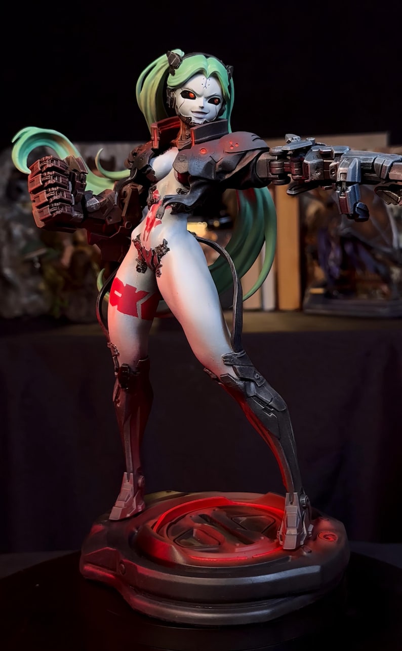 K&ouml;nnte beinhalten: Eine futuristische Figur einer weiblichen Figur mit gr&uuml;nen Haaren und einem wei&szlig;en Gesicht. Sie tr&auml;gt eine schwarz-rote R&uuml;stung und h&auml;lt eine gro&szlig;e Waffe. Die Figur steht auf einem kreisf&ouml;rmigen Sockel mit roter Beleuchtung.