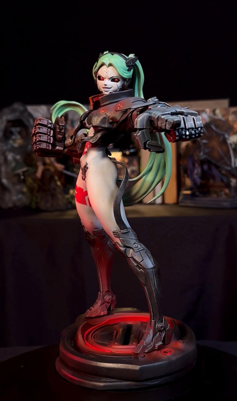 K&ouml;nnte beinhalten: Detaillierte Figur eines weiblichen Cyborgs mit gr&uuml;nen Z&ouml;pfen und roten Augen. Sie tr&auml;gt einen schwarz-grauen Panzeranzug mit roten Akzenten. Die Figur posiert mit geballten F&auml;usten auf einem kreisf&ouml;rmigen Sockel mit rotem Leuchten.
