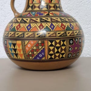 May include: A round ceramic pitcher with a handle, decorated with geometric patterns in brown, yellow, orange, green, and purple. The design is reminiscent of traditional pottery. The pitcher is a decorative item.