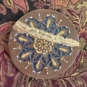 May include: A round, brown ceramic coaster with a blue and gold mandala design. The mandala features intricate details and a gold leaf accent.