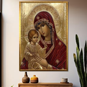 Puede incluir: Icono religioso enmarcado que representa a una madre y un niño en tonos dorados y rojos. La obra de arte tiene un fondo dorado en relieve y un marco dorado. La madre lleva una túnica roja y el niño túnicas doradas.