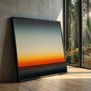 Puede incluir: Una impresión artística enmarcada que representa una puesta de sol sobre el océano. La obra de arte presenta un degradado de colores, incluyendo azul, naranja y amarillo. La impresión se muestra en un marco negro y se apoya contra una pared de hormigón sobre un suelo de madera.
