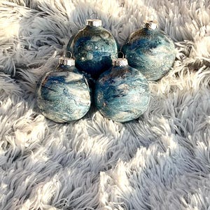 May include: Four glass Christmas ornaments with silver tops. The ornaments have a blue and white marbled design with silver glitter accents. They are arranged on a fluffy, white, faux fur surface.