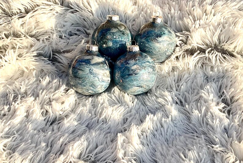 May include: Four glass Christmas ornaments with silver tops. The ornaments have a blue and white marbled design with silver glitter accents. They are arranged on a fluffy, white, faux fur surface.