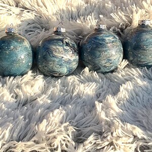 May include: Four glass Christmas ornaments with silver tops. The ornaments have a blue and silver marbled design with glitter accents. They are arranged on a fluffy, white surface.