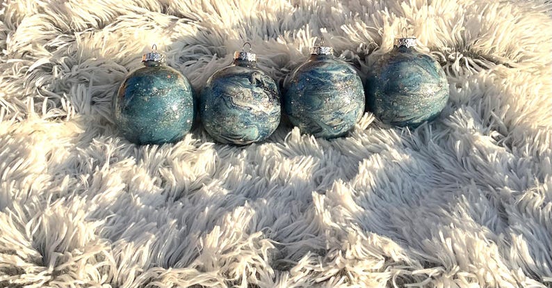 May include: Four glass Christmas ornaments with silver tops. The ornaments have a blue and silver marbled design with glitter accents. They are arranged on a fluffy, white surface.
