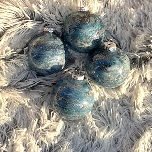 May include: Four blue and silver Christmas ornaments with a marbled effect. Each ornament has a silver cap and is displayed on a fluffy, white and gray surface. The ornaments have a shiny, glittery appearance.