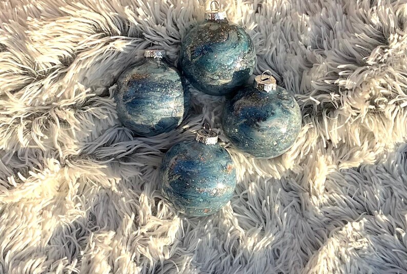 May include: Four blue and silver Christmas ornaments with a marbled effect. Each ornament has a silver cap and is displayed on a fluffy, white and gray surface. The ornaments have a shiny, glittery appearance.