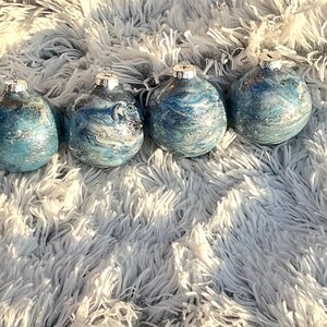 May include: Four blue and silver decorative ornaments with silver tops. The ornaments have a marbled effect and are arranged on a fluffy, white surface. The ornaments are teardrop-shaped.