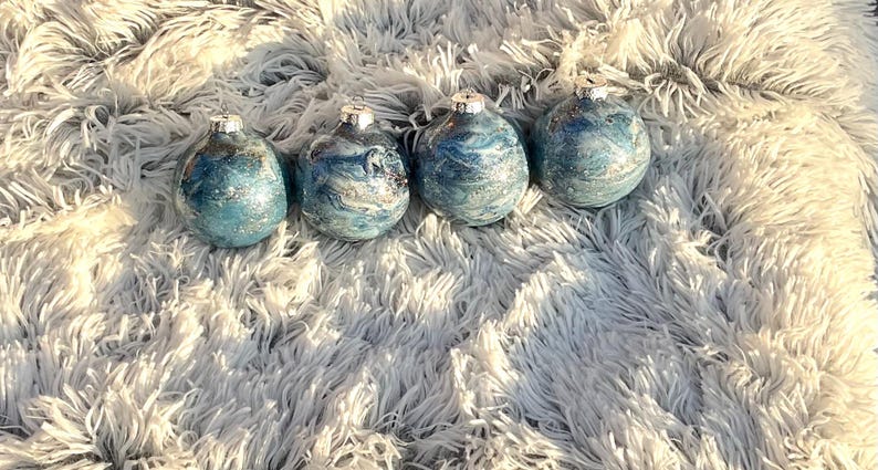May include: Four blue and silver decorative ornaments with silver tops. The ornaments have a marbled effect and are arranged on a fluffy, white surface. The ornaments are teardrop-shaped.