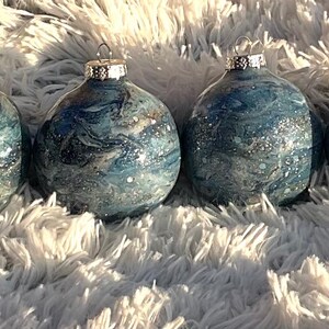 May include: Four blue and silver Christmas ornaments with a marbled effect. Each ornament has a silver cap and a loop for hanging. The ornaments are displayed on a fluffy, white surface.