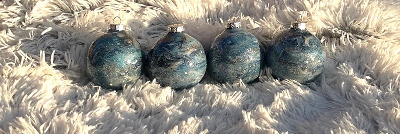 May include: Four blue and silver Christmas ornaments with a marbled effect. Each ornament has a silver cap and a loop for hanging. The ornaments are displayed on a fluffy, white surface.