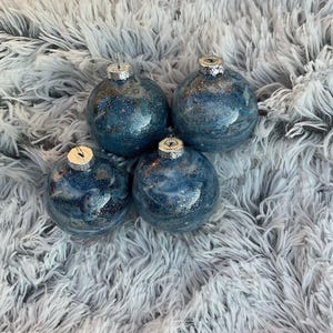 May include: Four blue and silver glitter Christmas ornaments with silver caps. The ornaments have a marbled effect and are sitting on a fluffy, white surface.