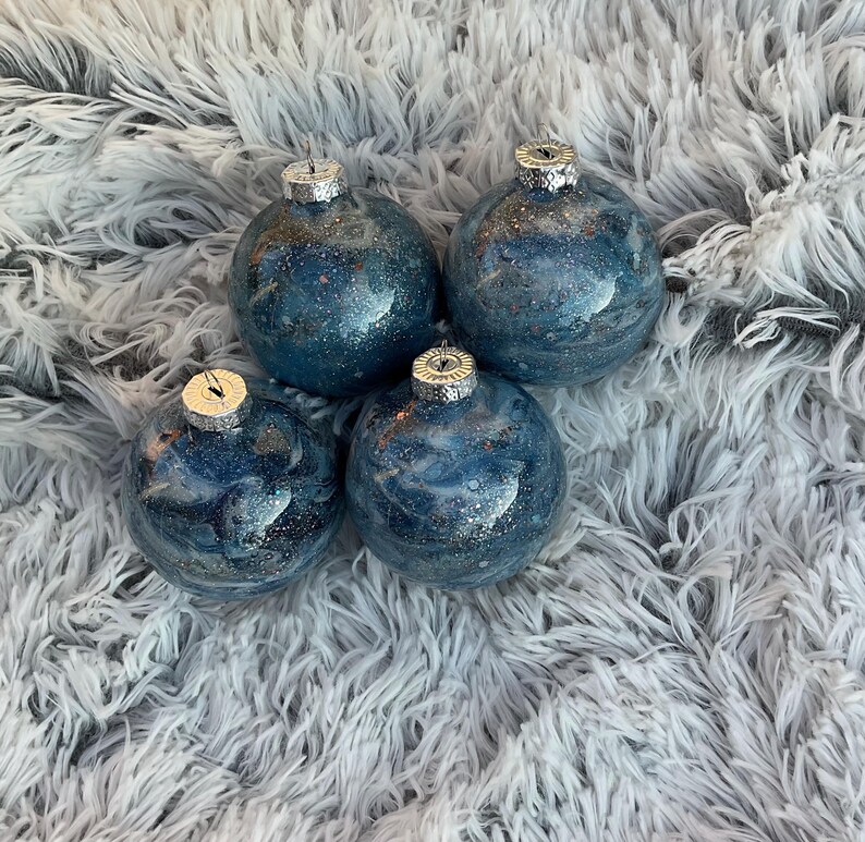 May include: Four blue and silver glitter Christmas ornaments with silver caps. The ornaments have a marbled effect and are sitting on a fluffy, white surface.