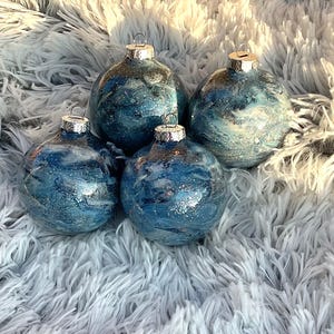 May include: Four blue and silver Christmas ornaments with a marbled effect. Each ornament has a silver cap and a loop for hanging. The ornaments are arranged on a fluffy, white surface.
