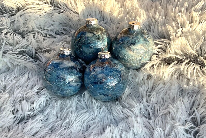 May include: Four blue and silver Christmas ornaments with a marbled effect. Each ornament has a silver cap and a loop for hanging. The ornaments are arranged on a fluffy, white surface.