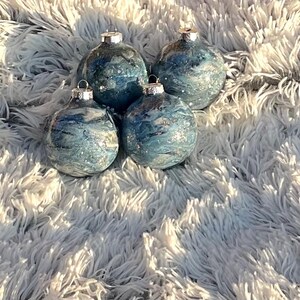 May include: Four blue and silver Christmas ornaments with a marbled design. The ornaments have silver tops and are arranged on a fluffy, white surface. The ornaments are round and have a shiny finish.