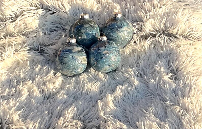 May include: Four blue and silver Christmas ornaments with a marbled design. The ornaments have silver tops and are arranged on a fluffy, white surface. The ornaments are round and have a shiny finish.