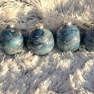May include: Four blue and silver marbled glass Christmas ornaments with silver caps. The ornaments are arranged in a row on a fluffy white surface. The ornaments have a swirling pattern of blue and silver.