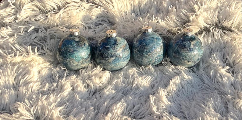 May include: Four blue and silver marbled glass Christmas ornaments with silver caps. The ornaments are arranged in a row on a fluffy white surface. The ornaments have a swirling pattern of blue and silver.