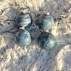 May include: Four blue and silver Christmas ornaments with a marbled effect. Each ornament has a silver cap and is displayed on a fluffy, white surface. The ornaments have a glittery, swirling design.