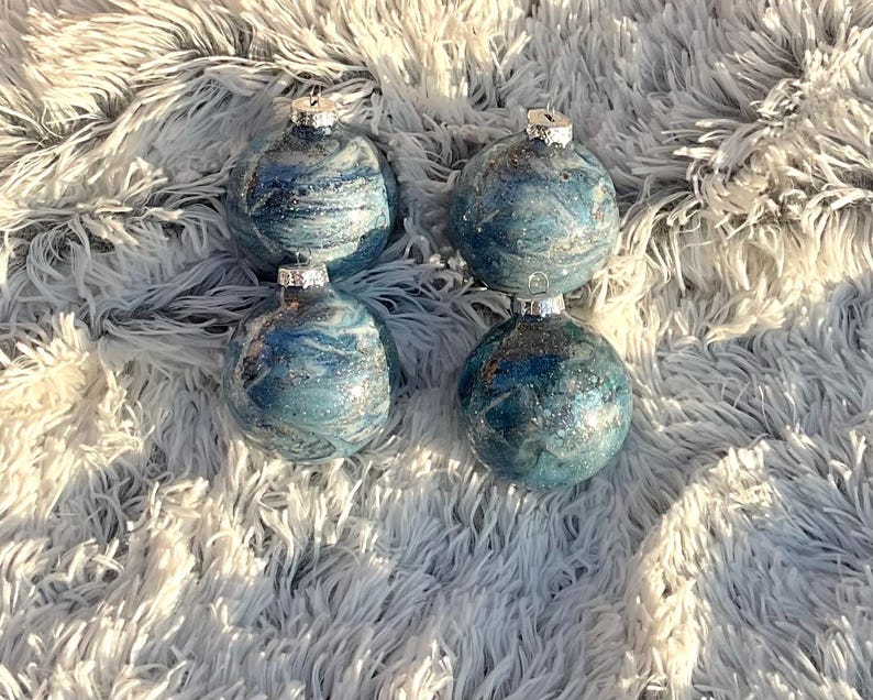 May include: Four blue and silver Christmas ornaments with a marbled effect. Each ornament has a silver cap and is displayed on a fluffy, white surface. The ornaments have a glittery, swirling design.