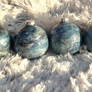 May include: Four round Christmas ornaments with a blue and silver marbled design. Each ornament has a silver cap and a loop for hanging. The ornaments are displayed on a fluffy, white surface.