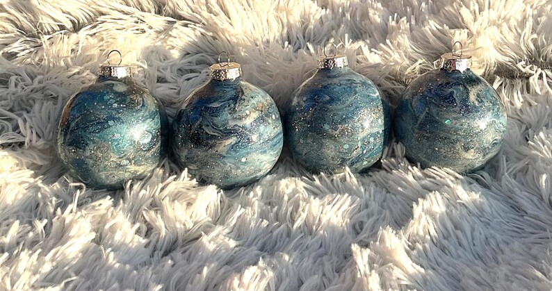 May include: Four round Christmas ornaments with a blue and silver marbled design. Each ornament has a silver cap and a loop for hanging. The ornaments are displayed on a fluffy, white surface.