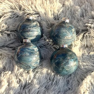 May include: Four round Christmas ornaments with a marbled blue and silver design. Each ornament has a silver cap and is displayed on a fluffy, white and gray surface. The ornaments are a festive decoration.