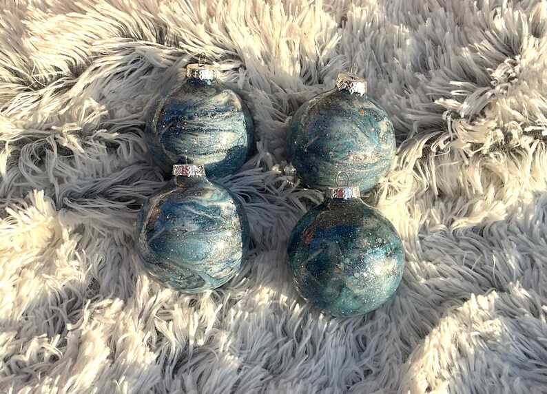 May include: Four round Christmas ornaments with a marbled blue and silver design. Each ornament has a silver cap and is displayed on a fluffy, white and gray surface. The ornaments are a festive decoration.