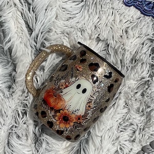May include: A glittery gold tumbler featuring a central graphic of a white ghost, an orange pumpkin, and autumn flowers, all surrounded by a black leopard print pattern. The mug includes a matching glittery gold handle and a black lid on top.