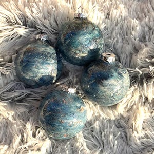 May include: Four blue and silver Christmas ornaments with silver metal caps. The ornaments have a marbled effect with silver glitter accents. They are arranged on a fluffy, white, faux fur surface.