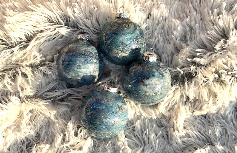 May include: Four blue and silver Christmas ornaments with silver metal caps. The ornaments have a marbled effect with silver glitter accents. They are arranged on a fluffy, white, faux fur surface.