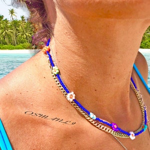 May include: A layered necklace featuring a blue beaded strand with flower accents, a gold chain, and a delicate silver chain. The necklace is adorned with colorful beads and small flower-shaped beads. The background shows a tropical beach scene.