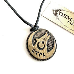 May include: A gold-toned pendant necklace with a black cord. The pendant features a wolf howling at the moon, a crescent moon, and a star. The pendant also has the word "AYDIN" inscribed on it.