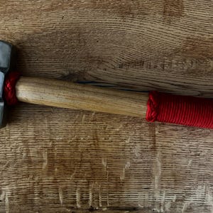 May include: A metal hammer with a wooden handle and a red wrapped grip. The hammer head has the text "DCI CHRISTIAN BARNES" and "FORGING THE FUTURE OF THE COAST GUARD" engraved on it. The hammer is lying on a wooden surface.