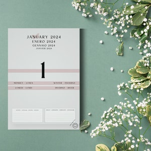 Puede incluir: Calendario de 2024 que muestra enero en varios idiomas. El diseño presenta un gran número 1, con secciones para notas. El calendario tiene una franja rosa pálido y está sobre un fondo verde suave con flores blancas.