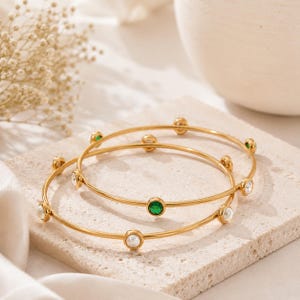 May include: Two thin gold-tone metal bangles featuring evenly spaced circular accents. Each bracelet is set with alternating round white pearls, clear crystals, and vibrant green gemstones, creating a delicate and symmetrical jewelry design.