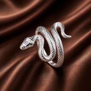 Puede incluir: Un anillo plateado con forma de serpiente enroscada que presenta un patrón texturizado de escamas en todo el cuerpo. La cabeza de la serpiente tiene una pequeña piedra verde brillante como ojo y la cola se curva hacia arriba en el diseño.