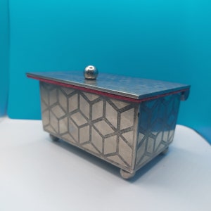 May include: A silver box with a geometric pattern and a red trim. The box has a round knob on the top.