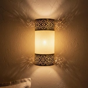 May include: A cylindrical wall sconce featuring a central off-white fabric shade flanked by two dark metal bands with intricate laser-cut patterns. The light emits a warm glow, casting decorative, symmetrical shadow patterns onto the wall surface.