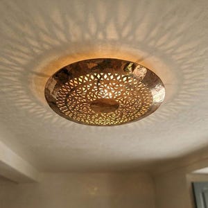 Peut inclure: Un plafonnier circulaire en laiton martelé doté d'un motif géométrique complexe découpé au laser. La lumière diffuse une lueur chaude à travers les découpes, projetant un motif d'ombre détaillé en forme d'étoile sur le plafond.