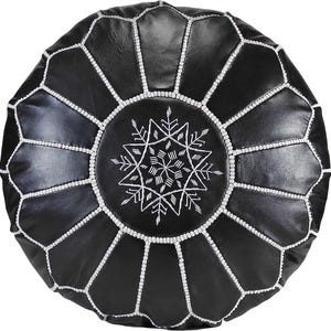 Peut inclure: Un pouf rond noir orné d'un motif d'étoile géométrique brodé en blanc au centre. La surface est divisée en douze panneaux par des coutures blanches rayonnantes partant du centre, offrant un aspect structuré et parfaitement symétrique.