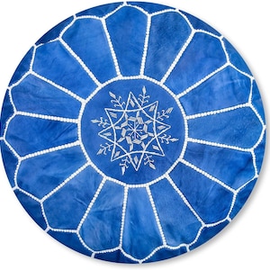 Peut inclure: Un pouf rond en cuir bleu vif orné d'un motif géométrique central en forme d'étoile brodé en blanc. La surface est divisée en douze panneaux en forme de pétales par des coutures blanches, créant un design radial distinct sur le dessus.