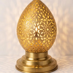 Peut inclure: Une lampe de table en métal doré en forme d'œuf, dotée d'un motif floral complexe découpé au laser qui laisse passer la lumière. La lampe repose sur une base circulaire de couleur laiton et émet une lueur chaude depuis l'intérieur.
