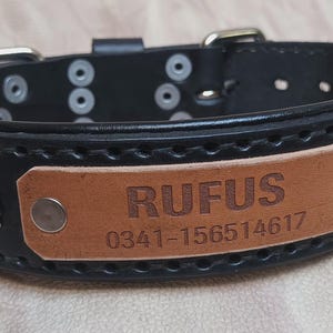 Może przedstawiać: Czarna sk&oacute;rzana obroża dla psa z brązową tabliczką z napisem "RUFUS" i numerem telefonu. Obroża ma srebrne metalowe akcenty i zapięcie na klamrę. Obroża ma szerokość około 3,8 cm.