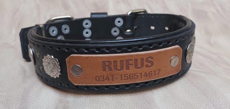 Może przedstawiać: Czarna sk&oacute;rzana obroża dla psa z brązową tabliczką z napisem "RUFUS" i numerem telefonu. Obroża ma srebrne metalowe akcenty i zapięcie na klamrę. Obroża ma szerokość około 3,8 cm.