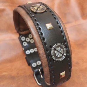 K&ouml;nnte beinhalten: Ein schwarzes Lederhundekragen mit braunem Lederbesatz. Das Halsband hat silberne und goldene Metallakzente, darunter ein Kleeblatt-Design und ein quadratischer Niet.