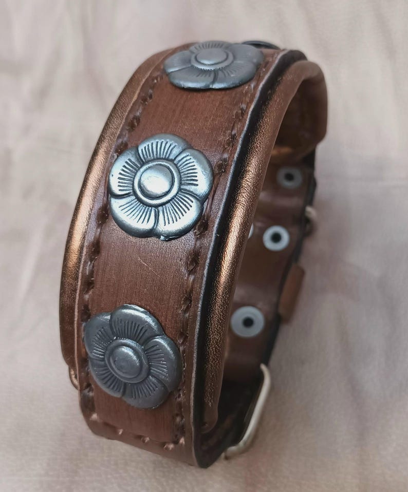 May include: A brown leather dog collar with silver flower-shaped accents. The collar has a buckle closure and is decorated with stitching.