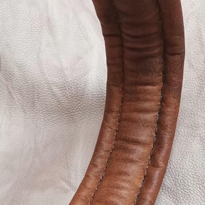 May include: Close-up of a curved, brown leather object with visible stitching. The leather has a textured appearance with darker areas, suggesting wear. The object is set against a white, textured background.